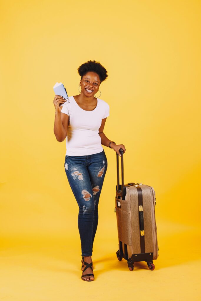 Smiling woman in casual attire with suitcase and passport on yellow background, ready for travel.