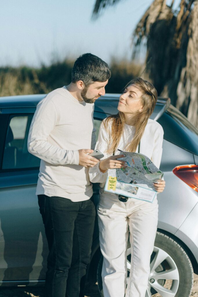 A young couple standing beside a car, holding a map and planning their road trip together outdoors.